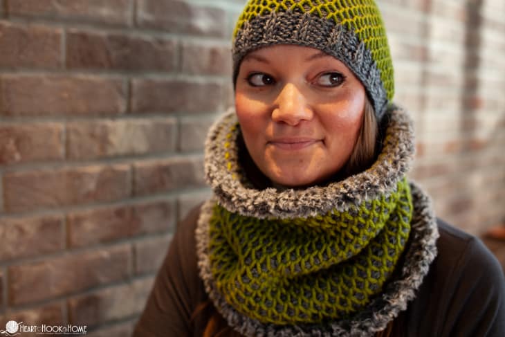 Tunisian “Smock it to Me” Crochet Set in Avocado and Cobblestone Heather colorways of Brava worsted and bulky yarn on a smiling female model by a brick wall.
