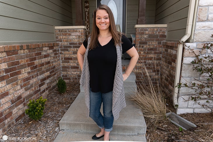 Smiling female model wearing an example of a light gray Spring Vest crochet pattern over a black shirt and jeans while standing in a brick entryway. 