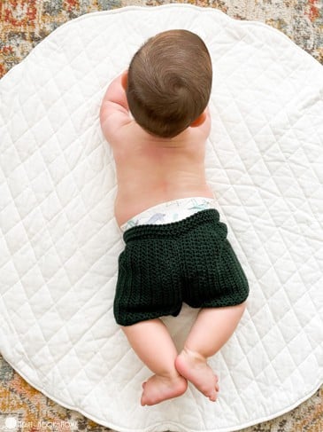 Infant model wearing an example of the Child Size Crochet Shorts in the “Ivy” colorway of Comfy Worsted yarn. The baby is laying on its stomach on a white circular rug. 