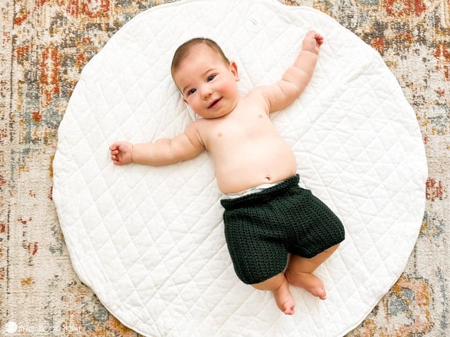 Infant model wearing an example of the Child Size Crochet Shorts in the “Ivy” colorway of Comfy Worsted yarn.  The baby is laying on its back on a white circular rug.  