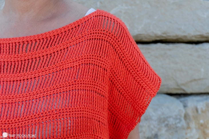 Shoulder view of the Track Stitch Tunic crochet pattern made with Shine Sport Yarn in the Grapefruit colorway on a female model standing in front of a stone wall.