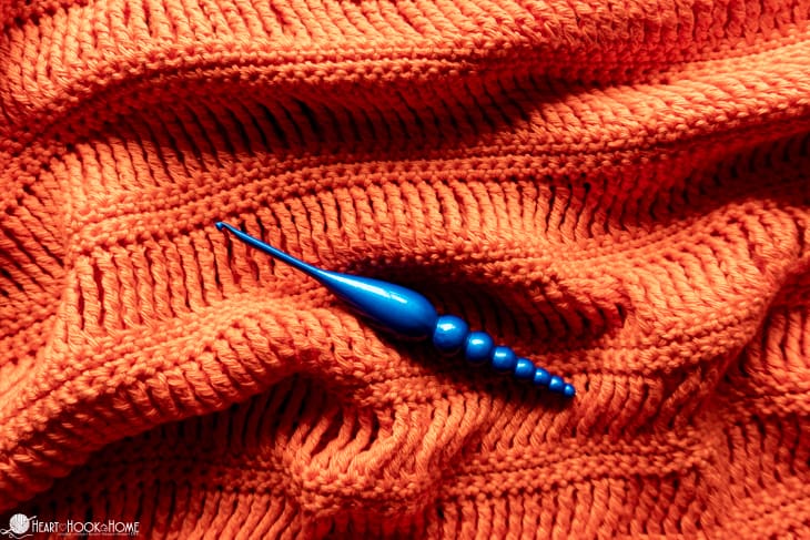 Sample of the Track Stitch Tunic crochet pattern made with Shine Sport Yarn in the Grapefruit colorway with a blue crochet hook laying in the center.