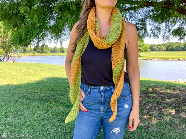 Green and Gold colored Tunisian Stardust Scarf crochet pattern on a female model wearing a dark blue tank top and distressed jeans in a park setting near a pond.  