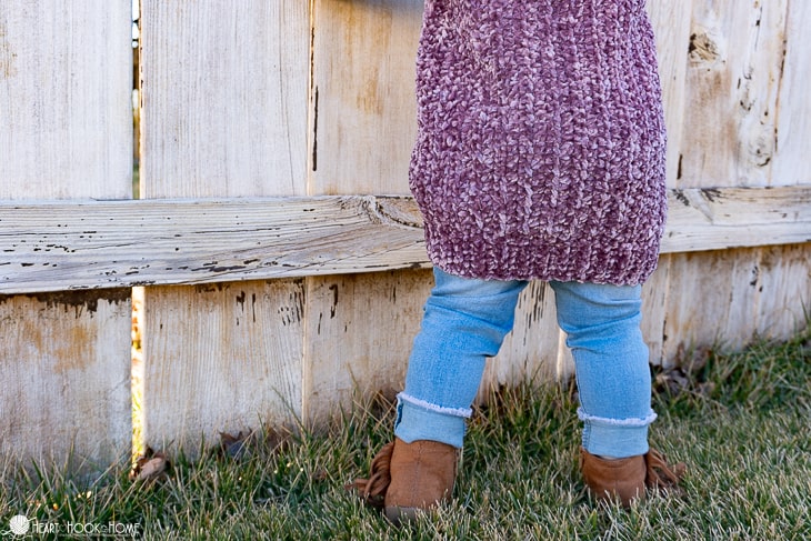 Purple colorway example of the Velvet Cardigan Crochet Pattern for Little Kids on a small child wearing jeans in a backyard setting.