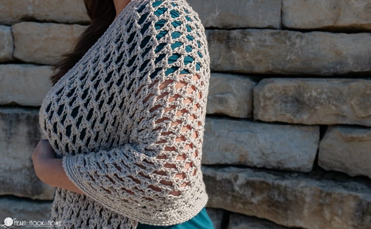 Ivory colored example of the Wanderlust Shawl crochet pattern on a female model standing near a brick wall.