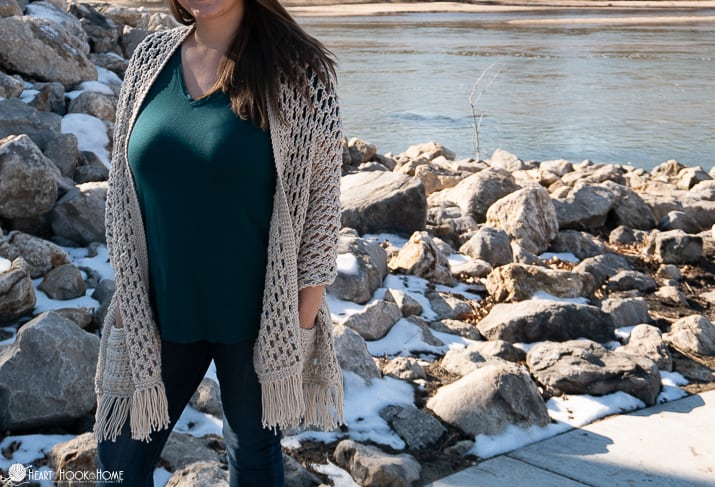Ivory colored example of the Wanderlust Shawl crochet pattern on a female model wearing a green shirt and standing near snow flecked rocks and a pond.