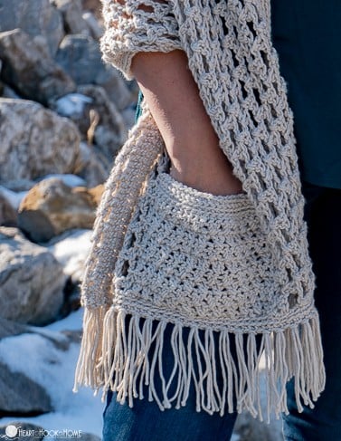 Ivory colored example of the pocket portion of the Wanderlust Shawl crochet pattern with a model\'s hand inserted inside and standing near snow flecked rocks.