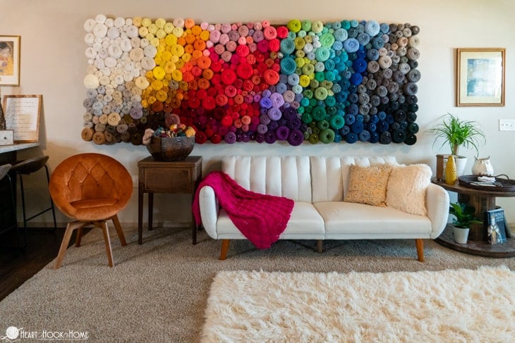 Yarn wall in an array of colors with a white couch with tan pillows and a Sangria Sorbet crochet blanket draped over it in a living rom with a furry, white rug over a gray carpet.