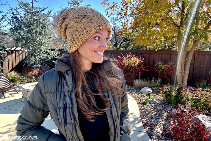 Gold example of the Back in Brugge Slouchy Beanie crochet pattern on a smiling female model wearing a brown jacket and black shirt in a backyard setting.
