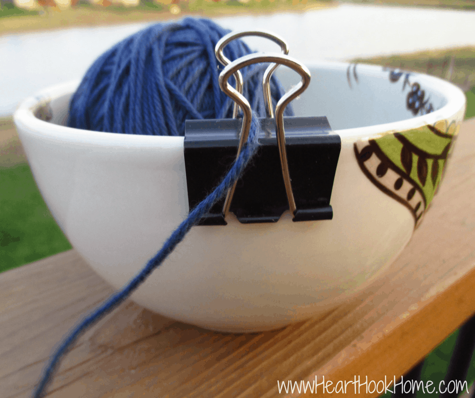 Blue ball of yarn in a decorative bowl with the tail of yarn being fed through a binder clip as a yarn guide.