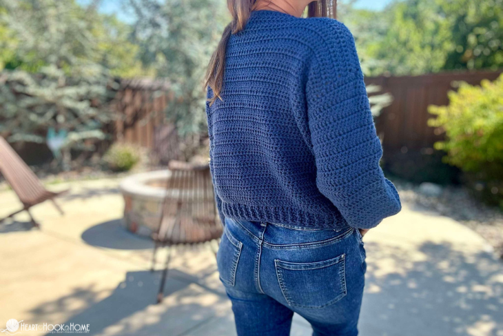 Back of a blue colorway example of a Blanche V-Neck Sweater crochet pattern on a female wearing blue jeans in a backyard setting.