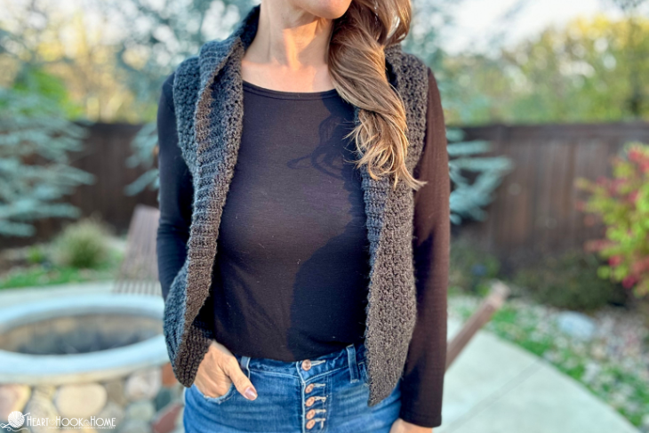 Gray example of the Layering Hooded Vest crochet pattern on a female model wearing a black, long sleeved shirt and jeans in a backyard setting.