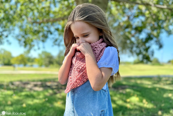 Female child wearing a light blue denim dress and a Lion Brand Coboo yarn in the “mauve” colorway example of the Frankie Cowl crochet pattern in a park setting.