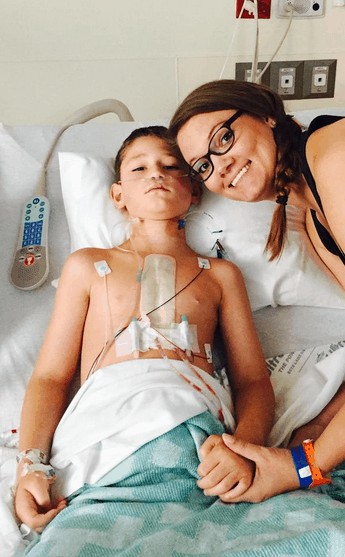 Smiling mom next to her son in a hospital bed who is post heart surgery and hooked up to medical monitors.