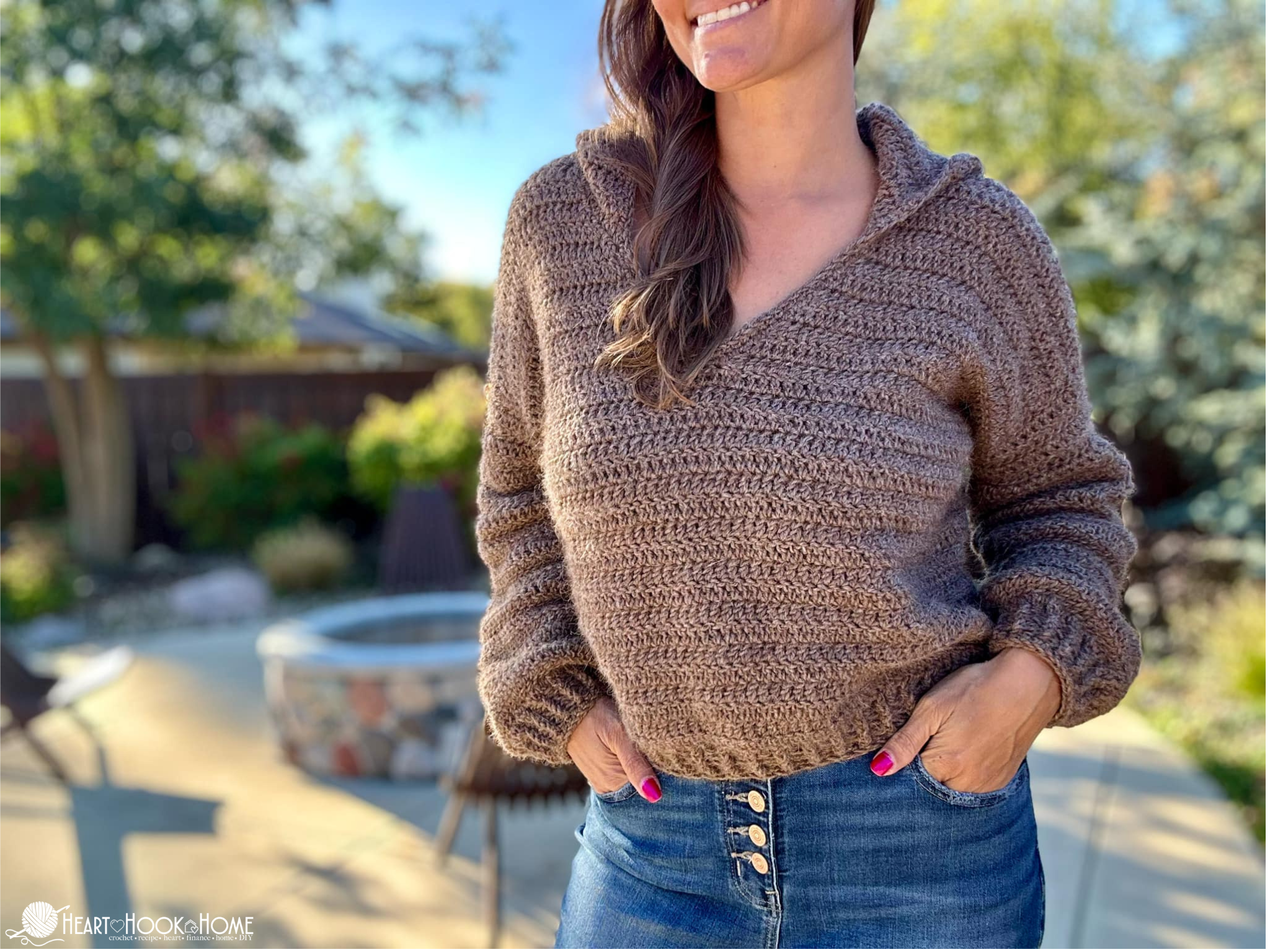 Light brown example of the Blanche V-Neck Sweater Crochet Pattern on a smiling female model wearing jeans in a backyard setting.